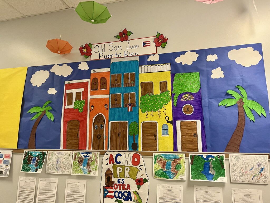 Classroom wall displaying a colorful mural of houses and palm trees. A banner reads "Old San Juan, Puerto Rico." Various artwork, including maps and drawings, is pinned below.