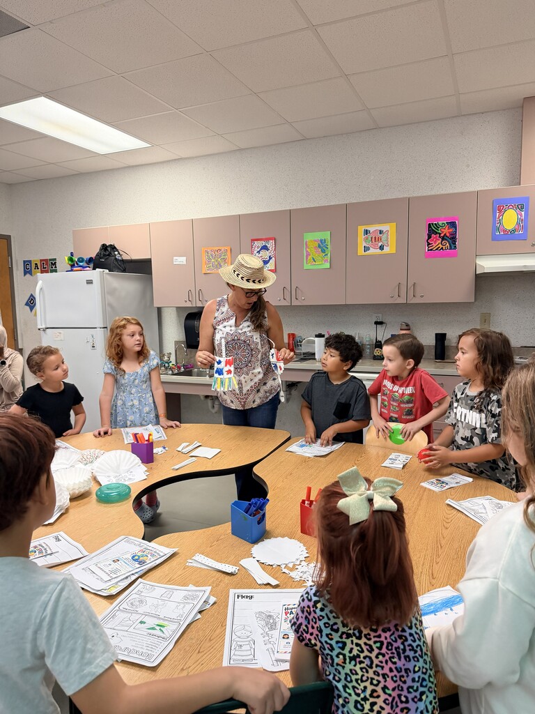 Children seated around a table with a teacher in a classroom, holding materials for an art project.