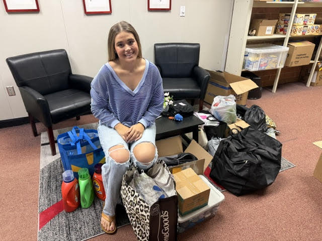 A woman in a blue top and ripped jeans sitting on the floor amidst various items.