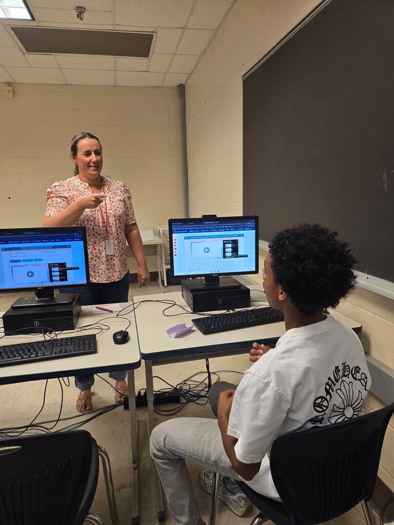A woman stands next to a desk with two computer monitors, talking to a student seated at the desk.