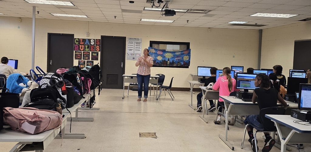 A classroom with a teacher standing. Students sit at desks with backpacks and computers.