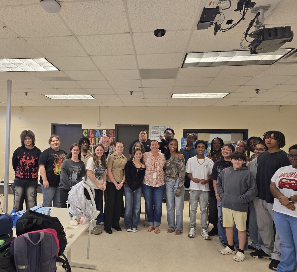Group of students and educators standing in a classroom. Some wear glasses, sneakers, and backpacks.