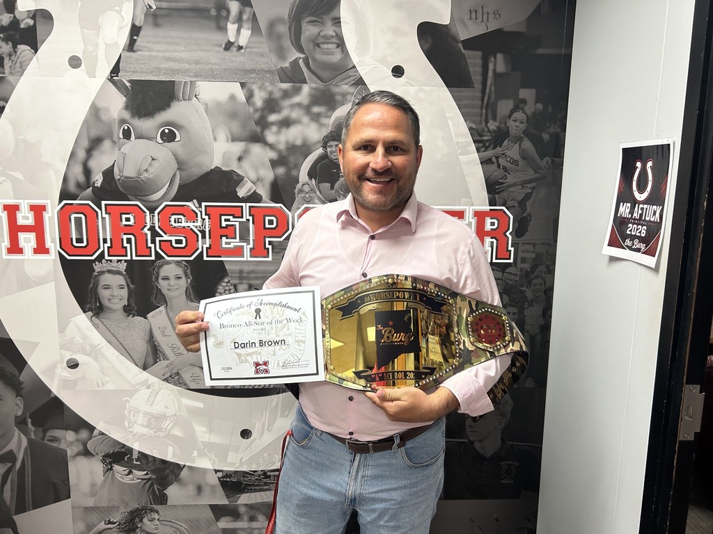 A man wearing a pink shirt and jeans holds a championship belt and certificate. Behind him is a large image of a horseshoe.