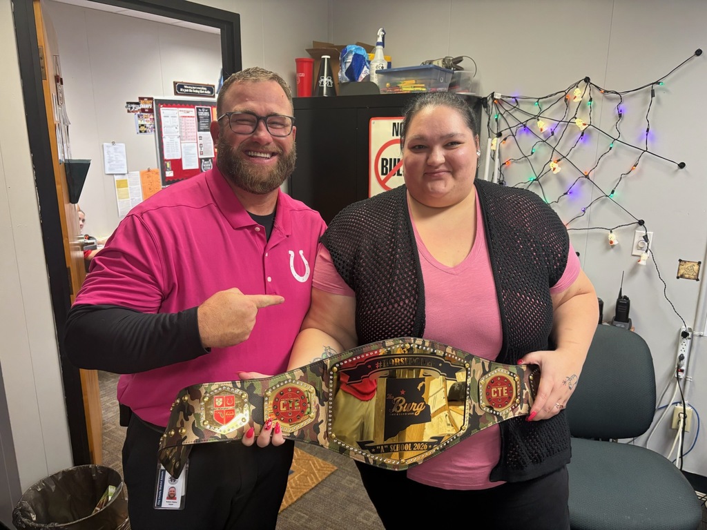 Two people standing and holding a championship belt. They stand in front of a wall with Christmas lights.