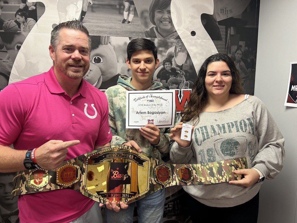 A man and two others hold a championship belt and certificate. A large, decorative wall is behind them.