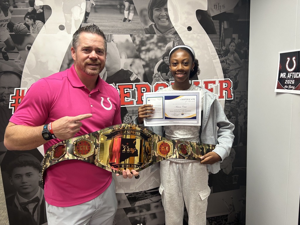 Man in pink shirt and a woman in gray sweatshirt holding a certificate and a championship belt.