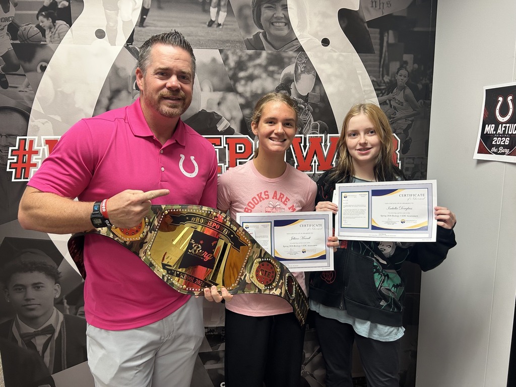Man in pink shirt and two girls hold belts and certificates, posing before a wall with sports memorabilia.