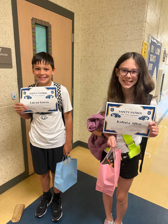 Two children standing, each holding a certificate and bags. A boy in white shirt and black shorts, a girl in white shirt and glasses.