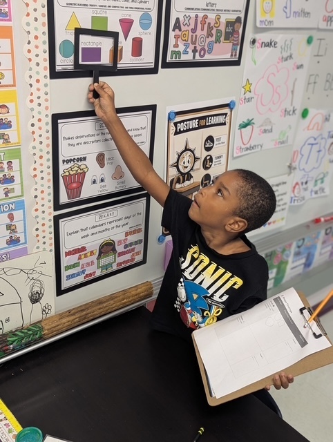 A young child with a clipboard holds a magnifying glass over a framed classroom bulletin board.