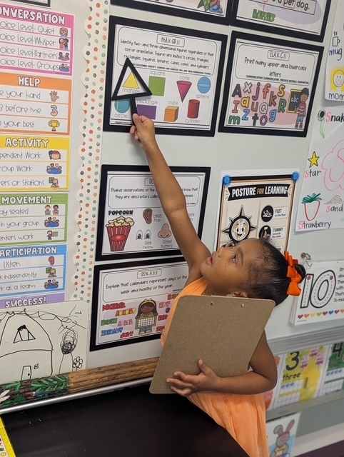 Child in orange dress with a bow touches a triangle shape on a classroom wall with educational posters.