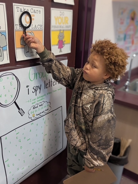 A child in camouflage clothing uses a magnifying glass to inspect a letter chart on a wall.