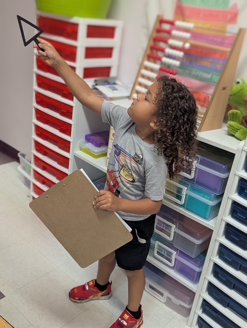 A child in a gray t-shirt and red shoes holds a triangular card and a folder. Shelves with colored storage boxes are behind.