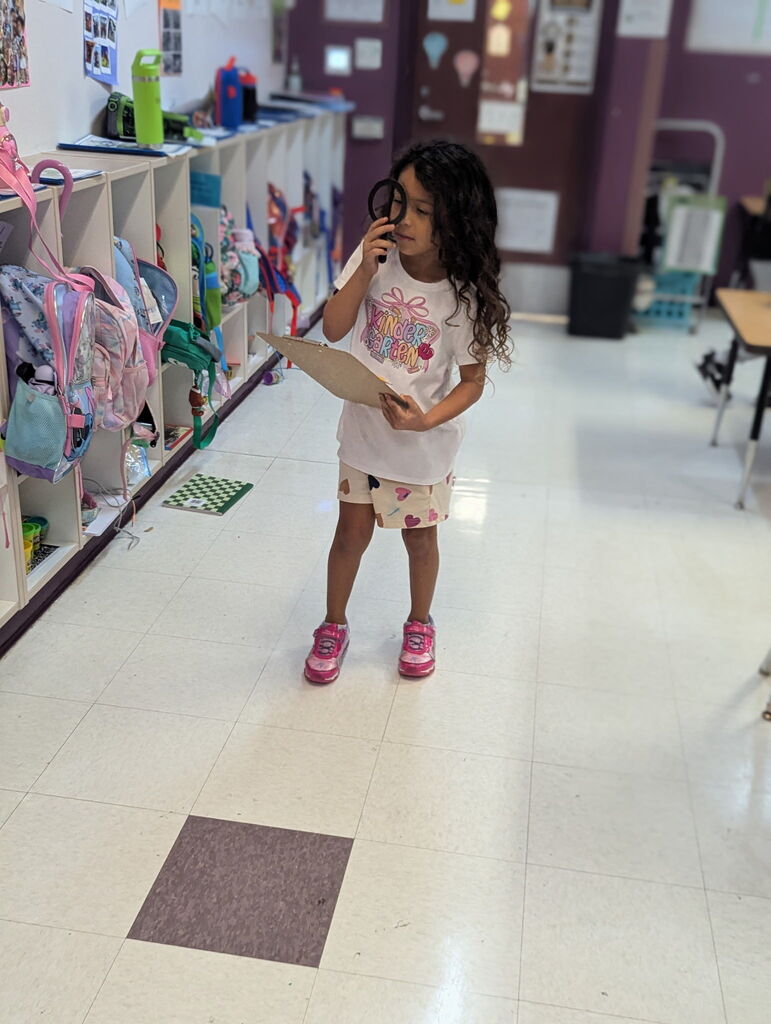 A young girl holds a magnifying glass to her eye and reads from a clipboard in a classroom hallway.