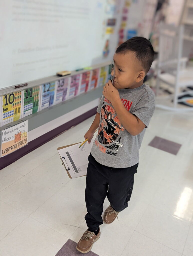 A young boy walks in a classroom, holding a clipboard and pen. He wears a gray shirt, dark pants, and brown shoes.