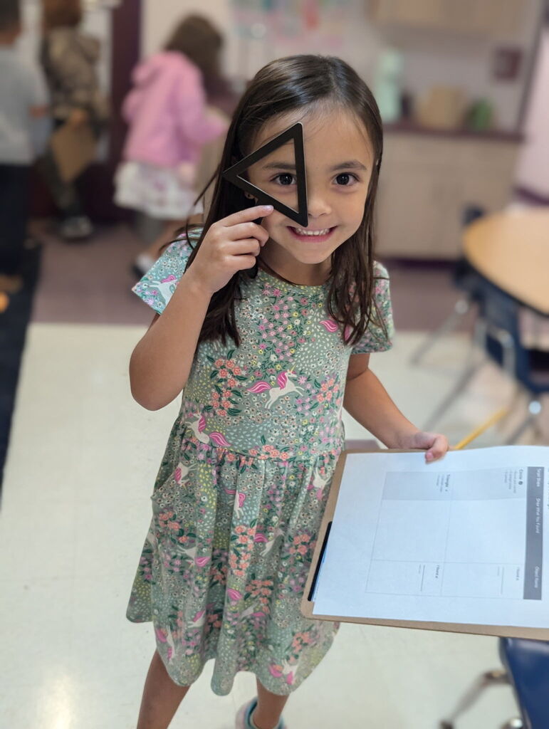 A girl holds a triangle-shaped object and a clipboard with paper. She smiles and looks at the camera. Other children are in the background.