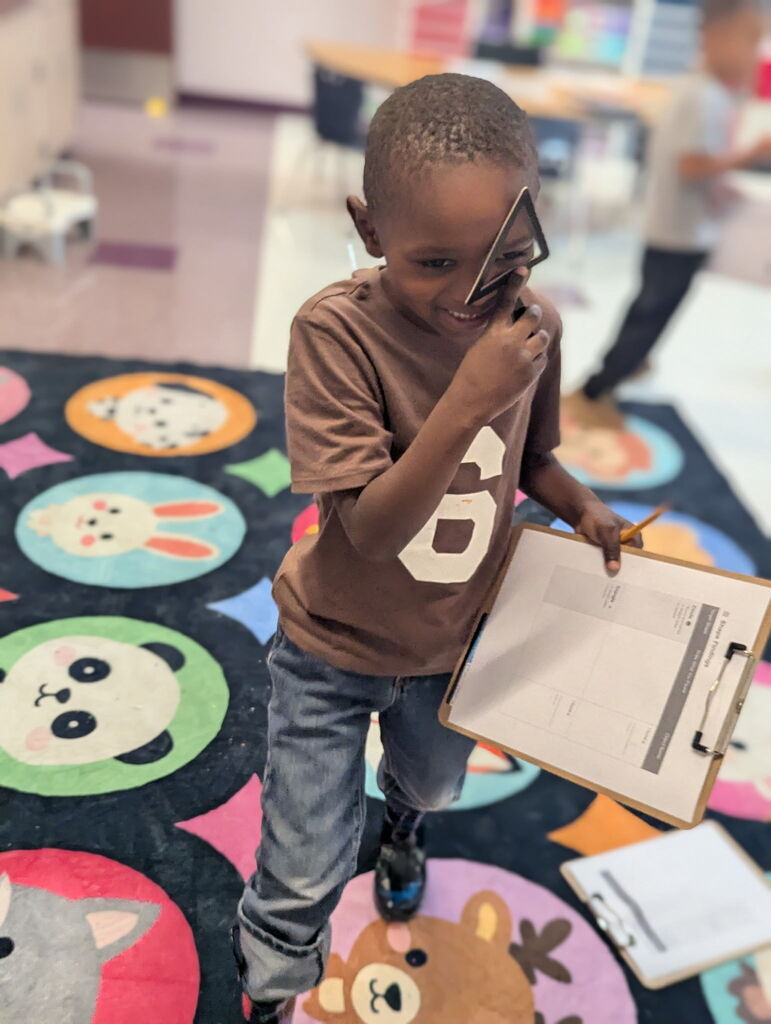 A child holds a clipboard with a pencil, wearing a brown shirt with number 8, standing on a multicolored rug.