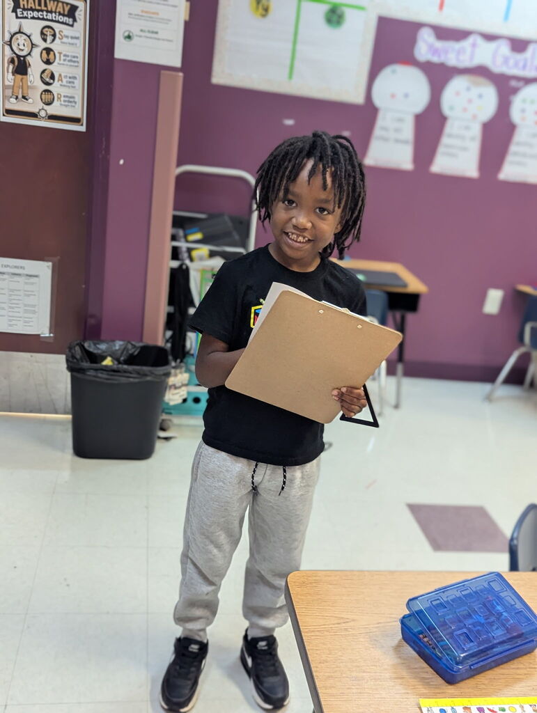 Child standing in classroom, holding a folder, smiling. Background shows purple walls, tables, and chairs.