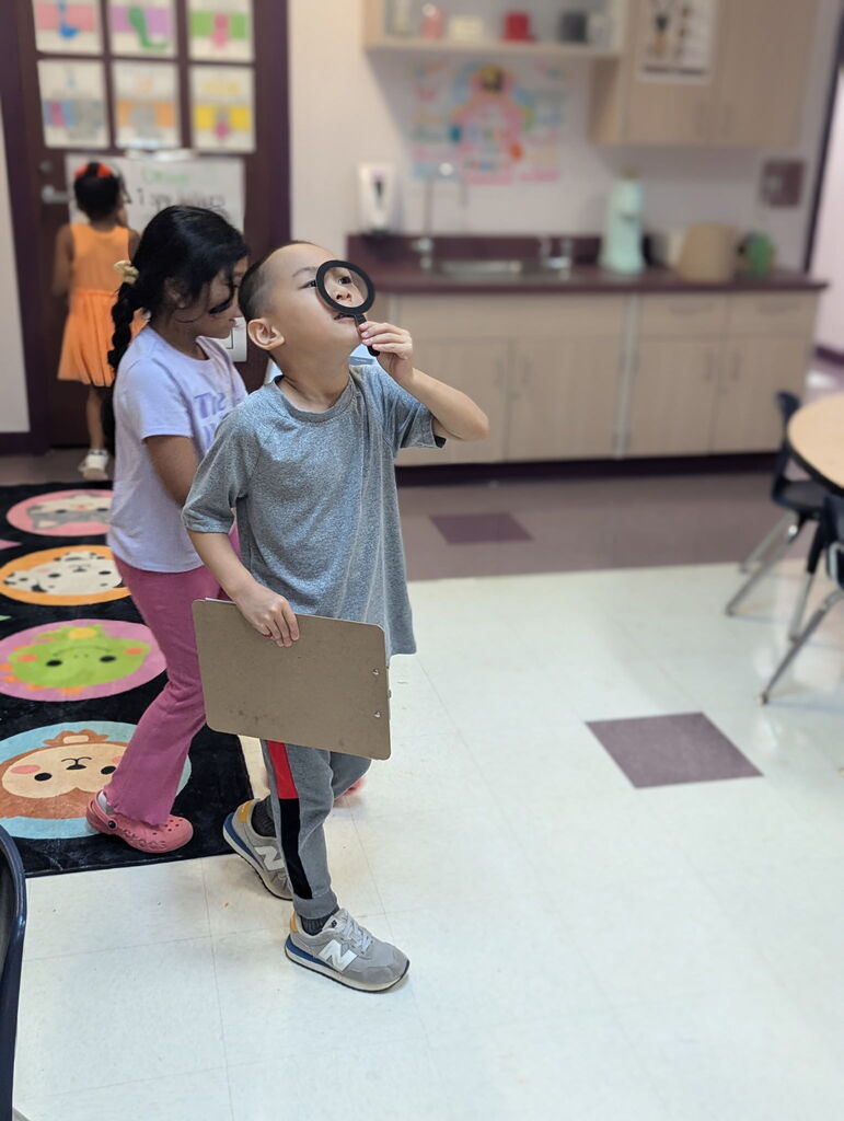 Two children observe an object with a magnifying glass in a classroom. One child holds a cardboard shield.