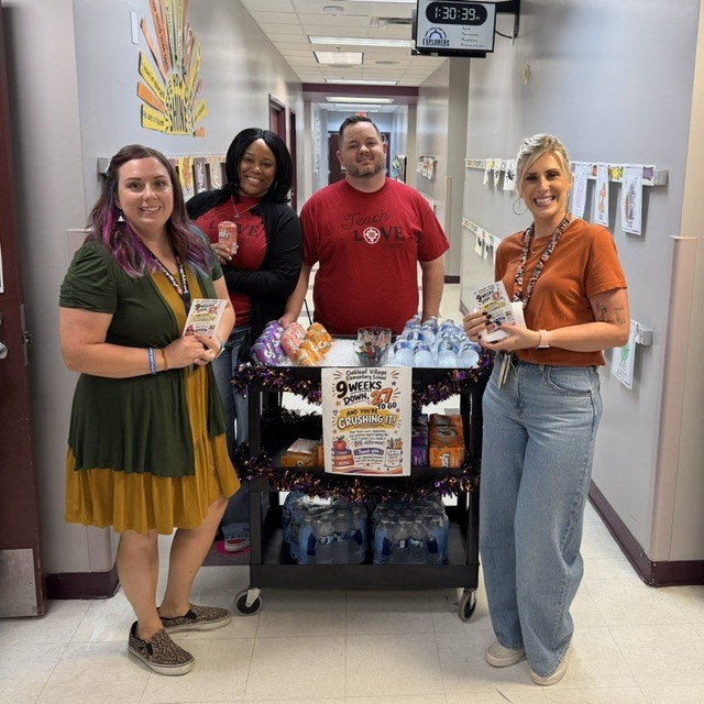 Four individuals pose with a cart of drinks and a poster in a hallway.