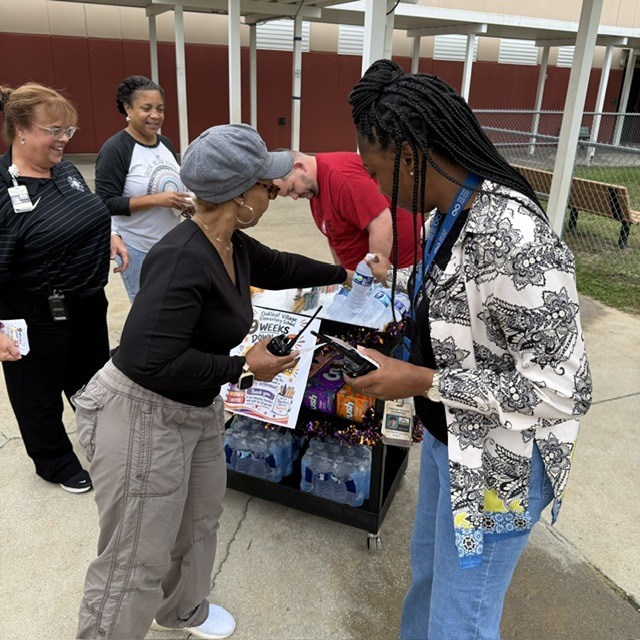 Adults stand outside, picking from a cart of drinks.