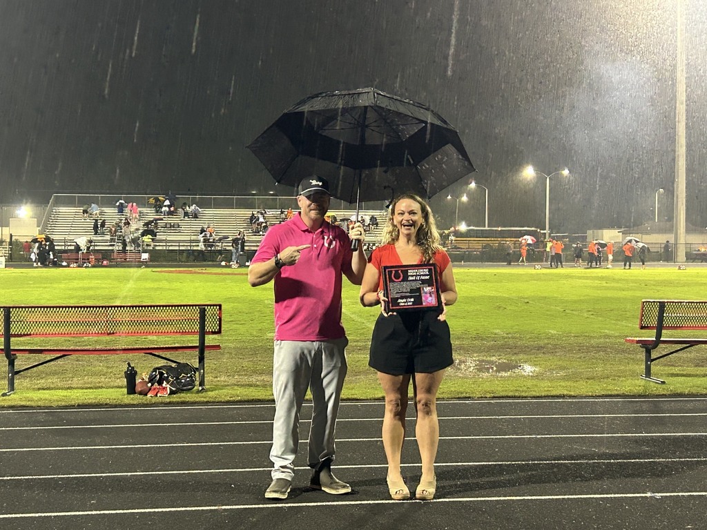A person holds an umbrella while standing with a woman in a pink shirt. They are on a track field.