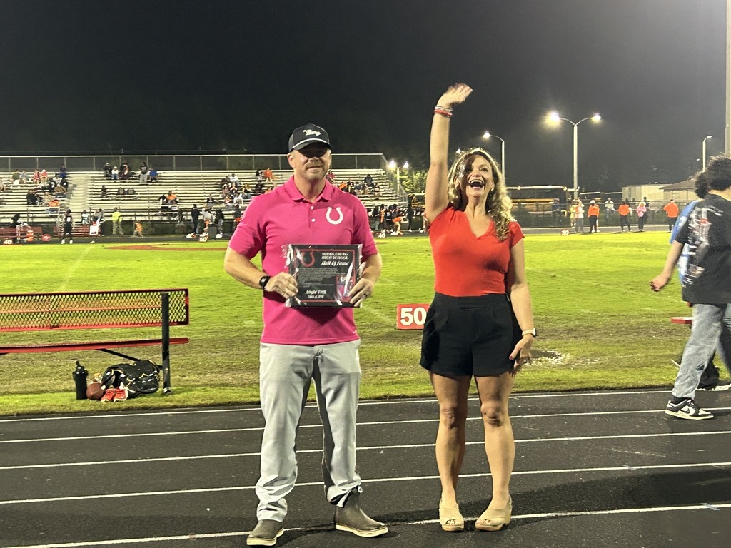 A man and a woman stand on a track at night. He holds a trophy. Behind them, people walk on the grass.