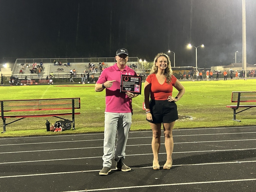 Two people, one in a pink shirt and the other in a red top, stand on a track holding an award.