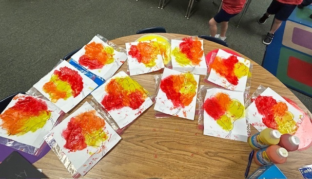 Table with circular artwork on papers. Bright colors, including yellow, orange, and red. Paint bottles on table.