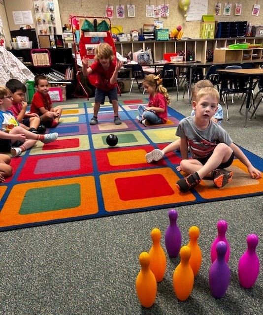 A group of young children in a classroom. Some are sitting on a colorful rug, others are standing. One child rolls a ball toward bowling pins.