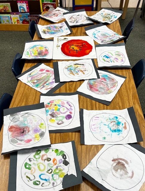 A table holds a row of circular artwork, each on a white paper with black borders. Background shows a bookshelf with books.