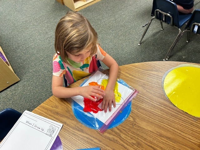 A young child is seated at a table, using paint on a paper sheet within a plastic bag.