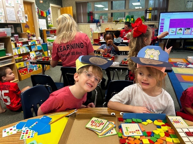 Children seated at a table in a classroom, with one wearing a hat, doing crafts. A monitor is visible behind them.