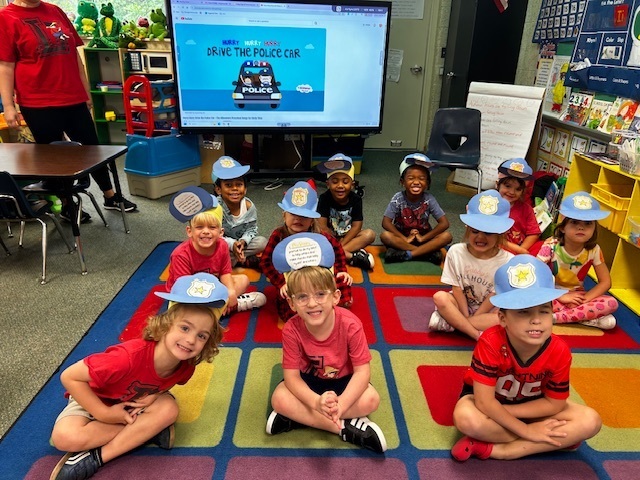 A classroom with children in hats sitting on a rug in front of a large screen.