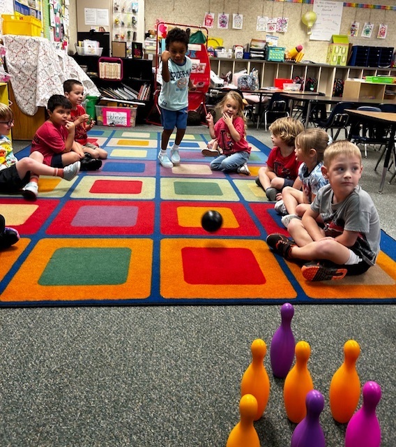 Children sit in a circle on a colorful rug, engaging in a game with bowling pins and a ball.