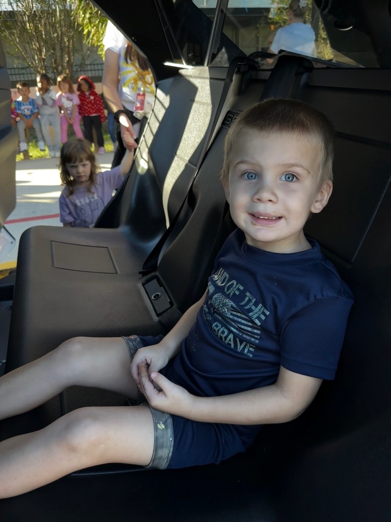 A young boy sits in the driver's seat of a fire truck with a safety belt. Behind him, a girl sits in the passenger seat. Several children wait outside.