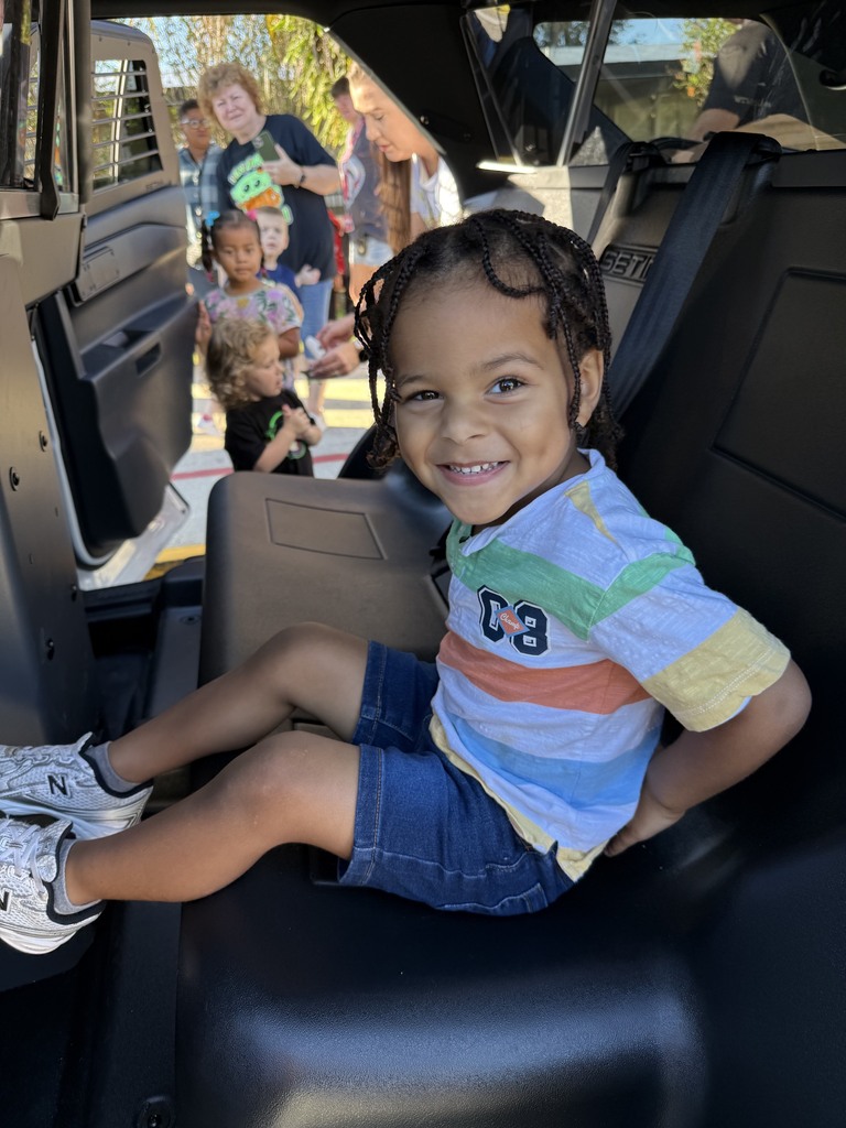 A child sits in a car seat, smiling, with other children visible in the background.