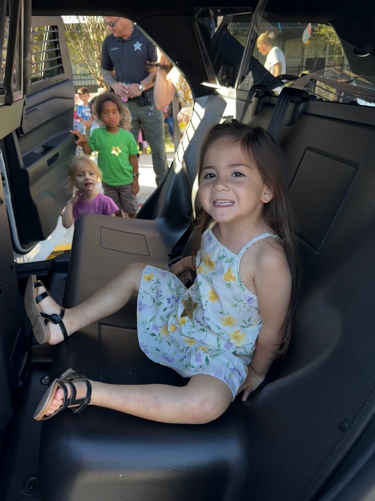 A smiling girl in a dress sits on a black car seat, with two children and a man in the background.