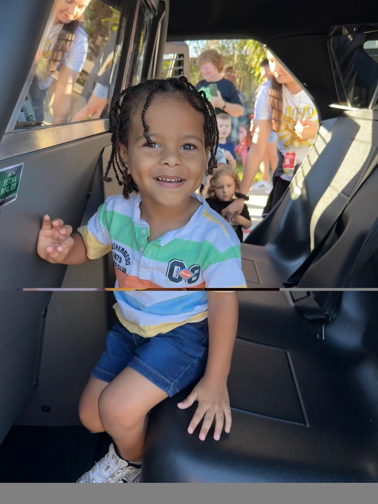 A smiling child sits inside a vehicle, wearing a striped shirt and denim shorts. Behind them, others are standing outside.