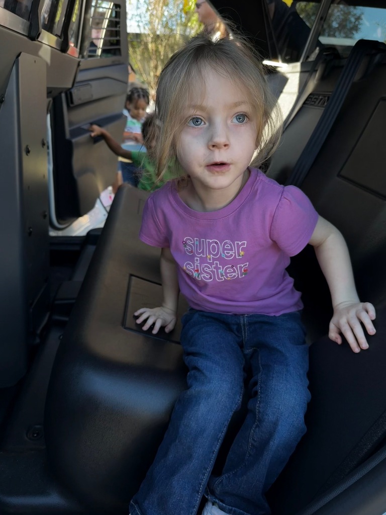 A young girl in a purple shirt with white letters sits in the back of a vehicle.