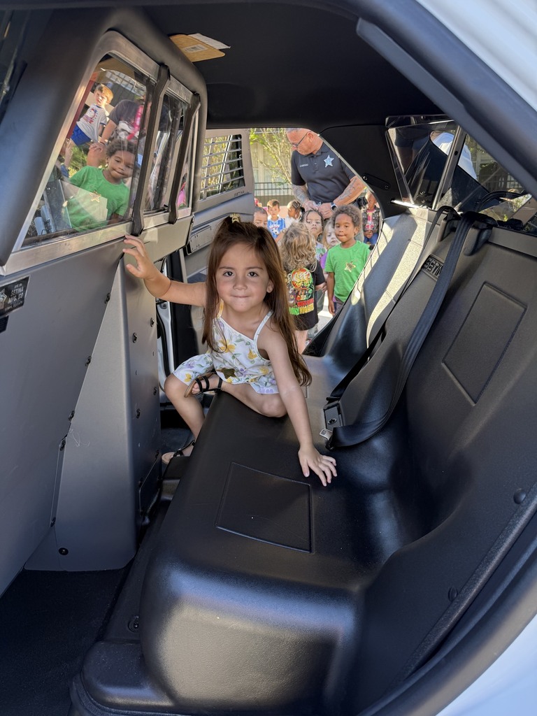 A young child sits in a police vehicle's back seat, with other children visible in the background.