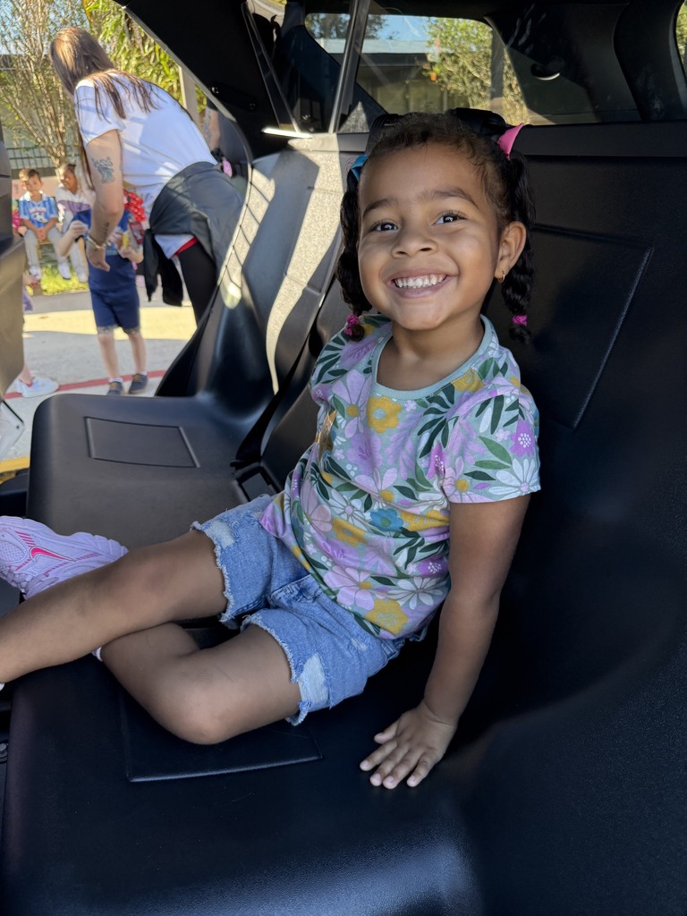 Child seated in a car's back seat, smiling. A person in the background is standing near another child.