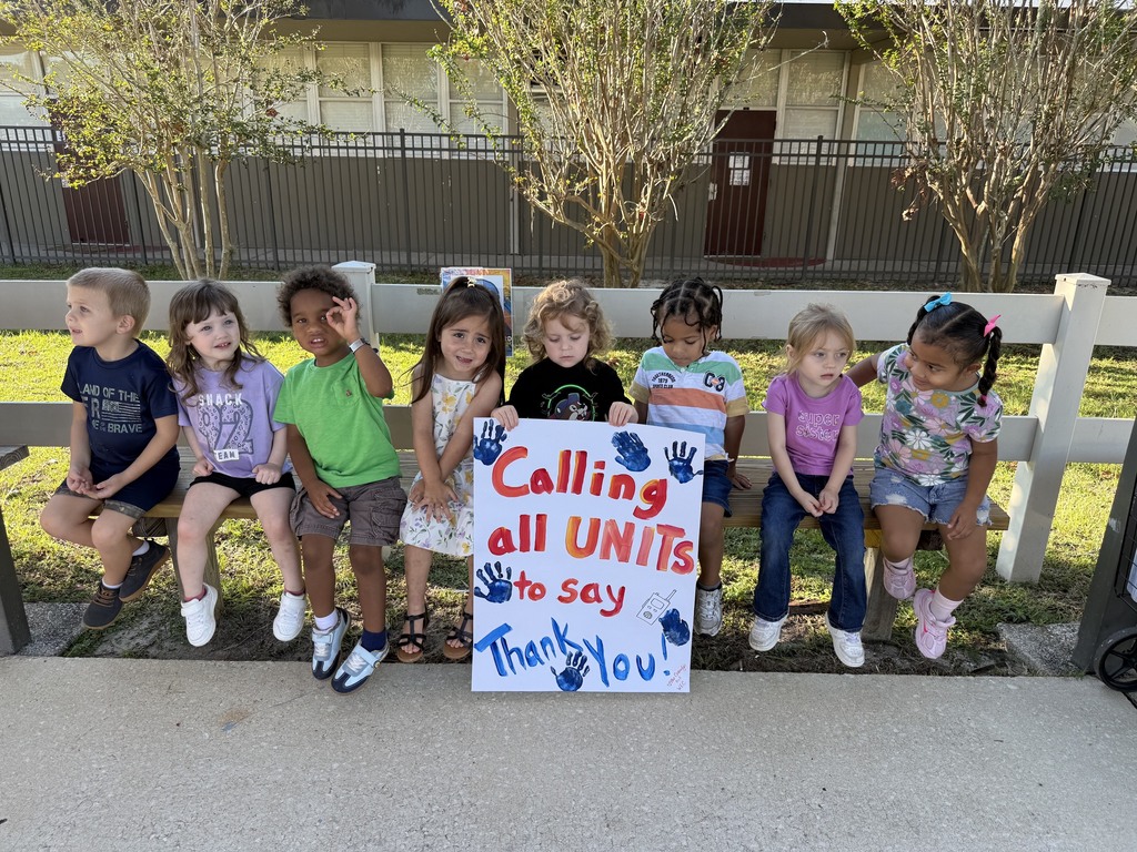 Nine children sitting on a bench with a sign that reads "Calling all units to say Thank You!" Trees and a building in the background.