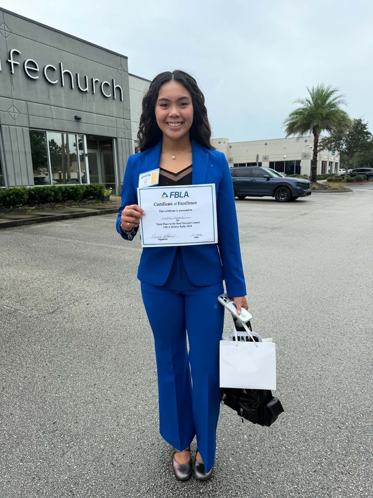 A woman stands in a parking lot, holding a certificate and shopping bags, dressed in a blue suit.