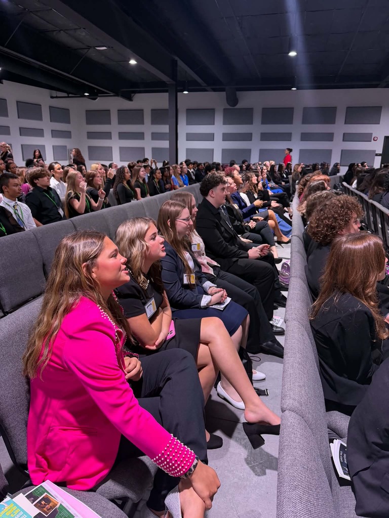 Many people seated in rows in a lecture hall; some wear name tags, and two wear pink and black coats.