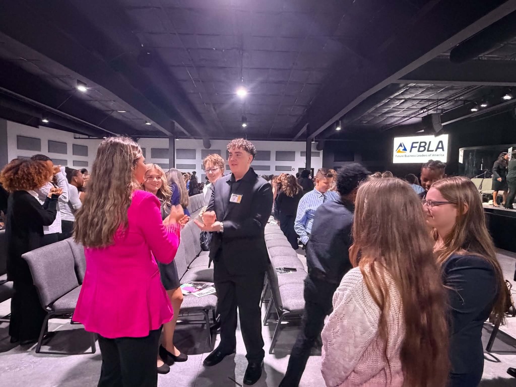 Several people dressed in formal wear standing and talking in a room with a yellow banner reading "FBLA".