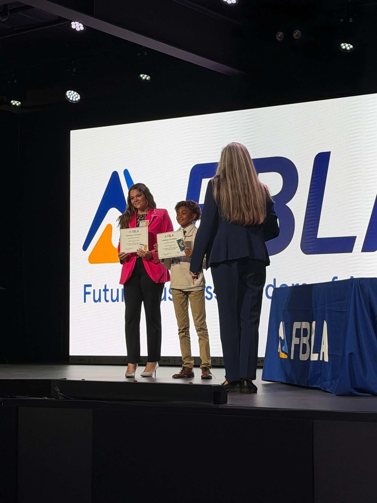 Three people on stage holding certificates. A large screen behind them displays "Future Students" with a logo.