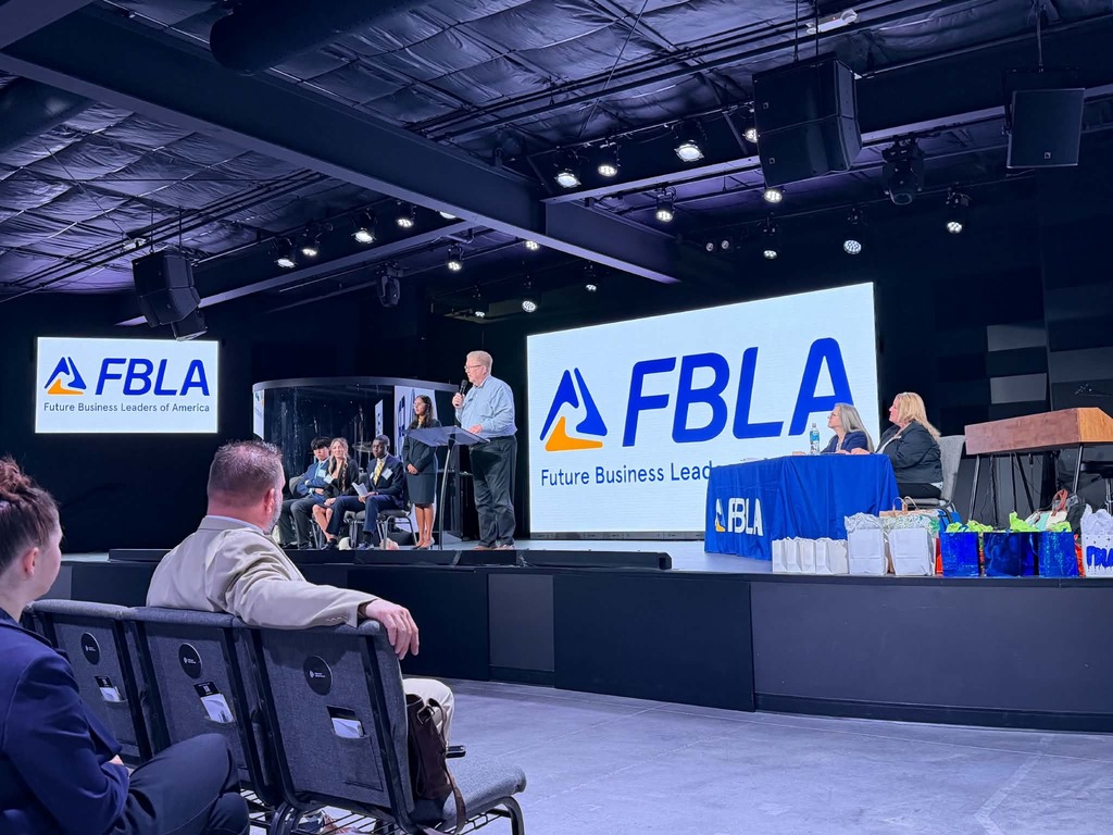 A conference room with a stage. A man speaks into a microphone. A large screen reads "FBLA".