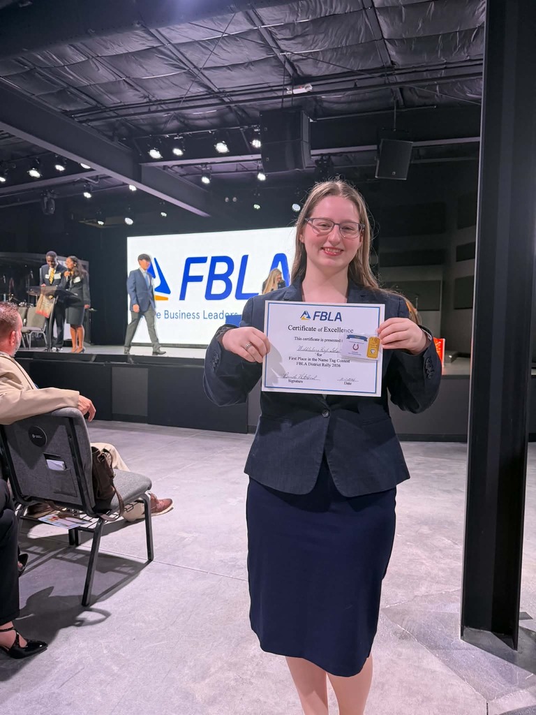 A woman holds a certificate and wears glasses and a blazer. Behind her is a stage with a screen reading "FBLA".