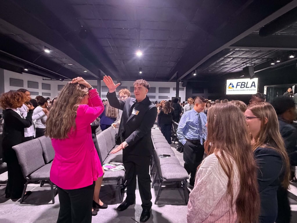 People in a room with chairs, a man in a black suit and woman in pink wave.