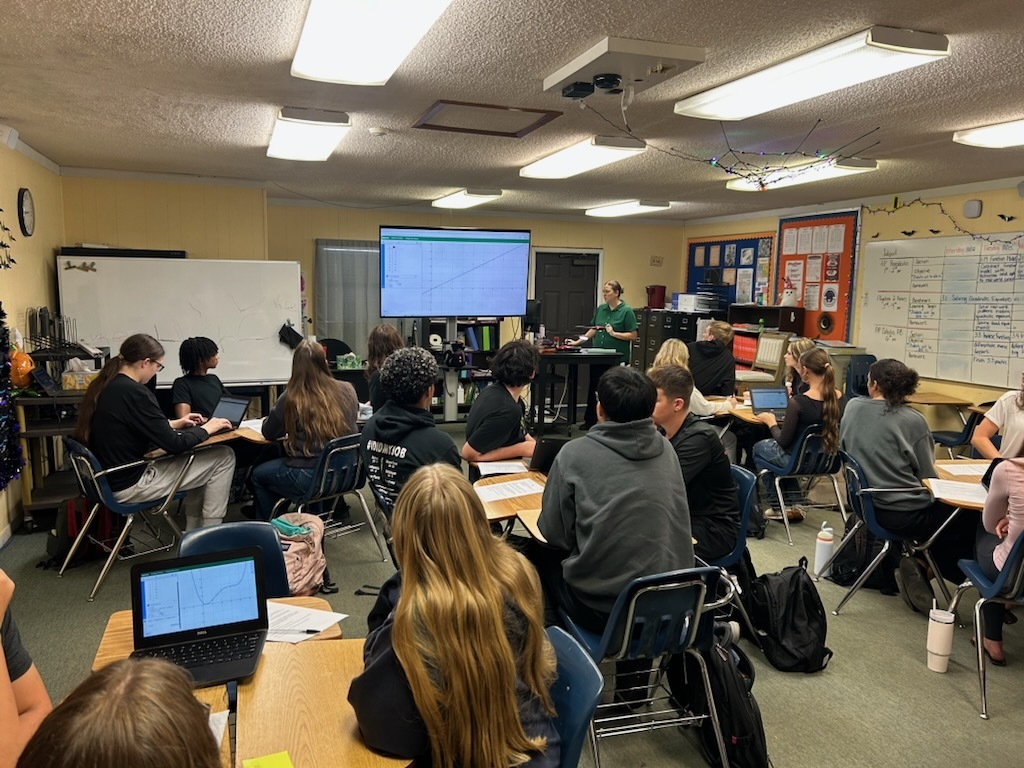 A classroom with students sitting at tables, laptops open, and a teacher near a projector screen.